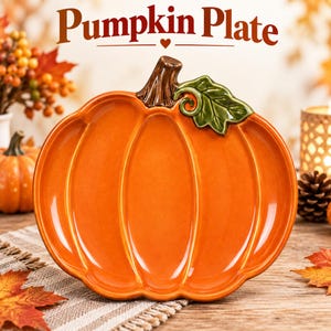 May include: A ceramic pumpkin-shaped plate with a glossy orange finish, a brown stem, and a green leaf accent. The text Pumpkin Plate is printed above the object in a dark brown serif font with a small heart icon centered below the text.