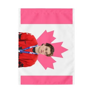 Könnte beinhalten: Eine rechteckige Gartenflagge mit weißem Hintergrund und einem großen rosa Ahornblatt in der Mitte. Eine Person in einem roten Sporttrikot mit schwarzem Nike-Logo und einem blauen Schlüsselband um den Hals ist über dem Ahornblatt abgebildet.