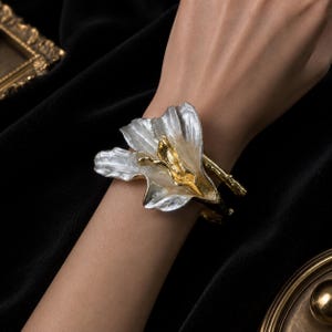 May include: A cuff bracelet featuring a large, sculptural flower design. The petals are finished in a textured silver tone, while the central stamen and the band are finished in a contrasting polished gold tone, creating a two-tone metallic look.