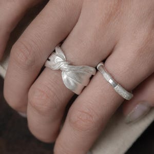 Könnte beinhalten: Zwei Silberringe an nebeneinanderliegenden Fingern. Ein Ring hat eine breite, mattierte Schiene, die zu einem Knoten geformt ist, der an eine Stoffschleife erinnert. Der zweite Ring ist eine schmale, strukturierte Schiene mit glatten und gekörnten Silbersegmenten.
