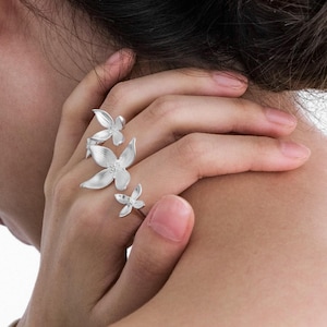 Könnte beinhalten: Ein silberner Ring mit drei unterschiedlich großen, blütenförmigen Elementen, die entlang der Ringschiene angeordnet sind. Die Blütenblätter haben eine matte, gebürstete Oberfläche mit kleinen, erhabenen kreisförmigen Details in der Mitte.