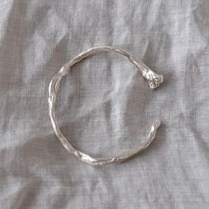 Könnte beinhalten: Ein silberfarbenes Armband in offener Form mit einer texturierten, organischen und unregelmäßigen Oberfläche, die an einen gedrehten Zweig oder Ast erinnert, mit leicht ausgestellten und unebenen Enden für eine markante, rohe Optik.