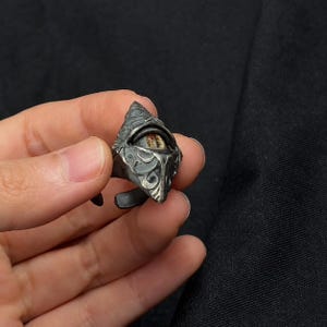 Könnte beinhalten: Ein Ring aus dunkel silberfarbenem Metall mit einem komplizierten, geprägten Schnörkelmuster und einem zentralen, realistisch wirkenden Auge mit brauner Iris und vertikaler Pupille. Der Ring hat ein offenes, verstellbares Design mit oxidiertem Finish.