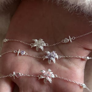 Könnte beinhalten: Drei silberfarbene Kettenarmbänder, jedes mit einem zentralen Blumenanhänger. Das obere Armband hat eine silberne Blume, das mittlere eine hellviolette Blume mit einem Herzanhänger und das untere eine hellrosa Blume mit einem Herz.