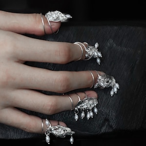 Könnte beinhalten: Ein Set aus fünf silberfarbenen Nagelringen mit texturierten, organisch geformten Metallkappen, die das Nagelbett bedecken. Jeder Ring ist mit kleinen, hängenden weißen Süßwasserperlen verziert und besitzt ein dünnes, verstellbares Silberband.