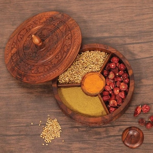 May include: A round wooden spice box with a carved lid, featuring four interior compartments filled with coriander seeds, turmeric powder, cumin powder, and dried red chili peppers. The container is crafted from dark brown wood with intricate patterns.