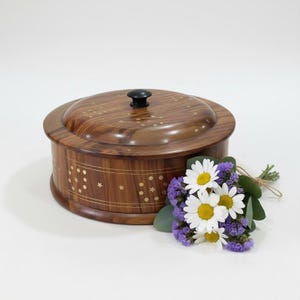 May include: A round wooden box with a polished brown finish and a dark knob on the lid. The exterior features inlaid brass patterns of small stars and dots arranged in circular motifs. A small bouquet of white daisies and purple flowers sits nearby.