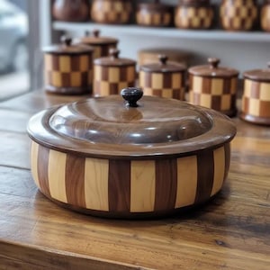 May include: A round wooden container with a lid, featuring alternating vertical stripes of dark walnut and light maple wood. The lid is smooth, dark brown, and topped with a small, centered black knob. The design is cylindrical and polished.
