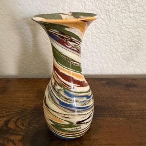May include: A small vase featuring a marbled pattern with swirling colours of cream, olive green, burnt orange, and deep blue. The vessel has a narrow neck that flares outward at the top rim, creating a smooth, rounded silhouette with a glossy finish.