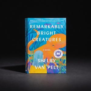 Op de afbeelding: Een boek getiteld Remarkably Bright Creatures van Shelby Van Pelt. De omslag toont een grote oranje octopus en een persoon met blauw haar tegen een blauwe onderwaterachtergrond met waterplanten. Een ronde badge staat rechtsonder.