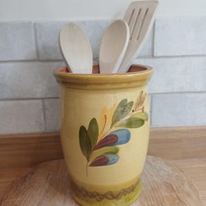 May include: A cylindrical yellow ceramic utensil holder featuring a hand-painted olive branch motif with green and blue leaves. The container holds two light-coloured wooden spoons and a wooden slotted spatula, showcasing a glazed finish.