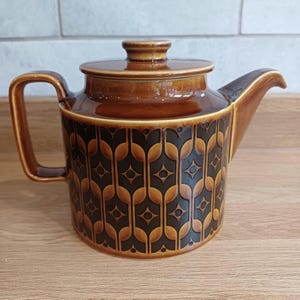 May include: A brown ceramic teapot with a glossy finish and a dark brown geometric pattern featuring repeating diamond and arch shapes. The teapot has a cylindrical body, a flat lid with a small knob, a curved handle, and a pointed spout.