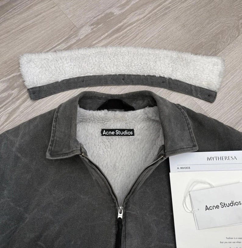 May include: A charcoal gray denim jacket with a white sherpa lining and a detachable sherpa collar. The jacket features a front zipper closure and an Acne Studios brand label on the inner neck. A white paper tag with the text Acne Studios is included.