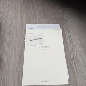 May include: A white rectangular document from Mytheresa featuring the text Mytheresa at the top and bottom, with an attached white Acne Studios brand tag. The document includes the text Fashion is a weapon that you use when you need it.