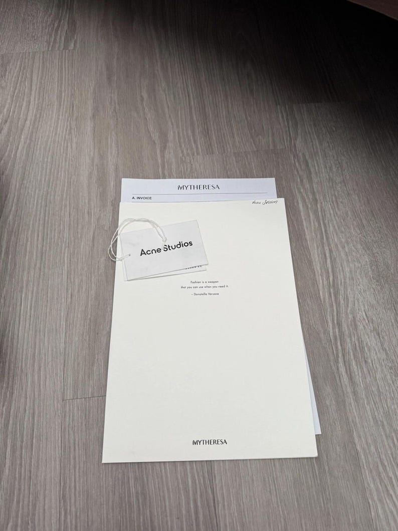May include: A white rectangular document from Mytheresa featuring the text Mytheresa at the top and bottom, with an attached white Acne Studios brand tag. The document includes the text Fashion is a weapon that you use when you need it.