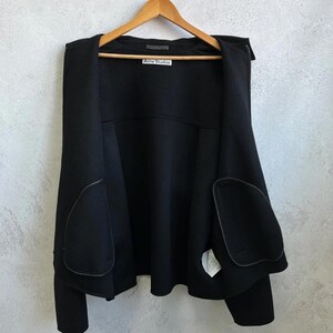May include: A black jacket hanging on a wooden hanger. The jacket features an open front design with two large, rounded patch pockets on the lower front panels. A white label with the text Acne Studios is stitched inside the neckline.
