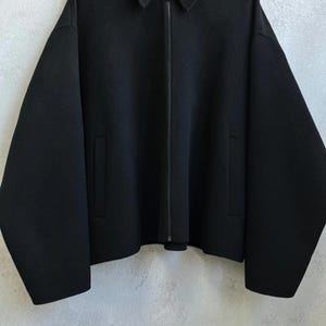 May include: A solid black jacket featuring a pointed collar, a full-length front zipper closure, and two vertical slit pockets. The garment has long, wide sleeves and a straight hemline, constructed from a smooth, dense fabric material.