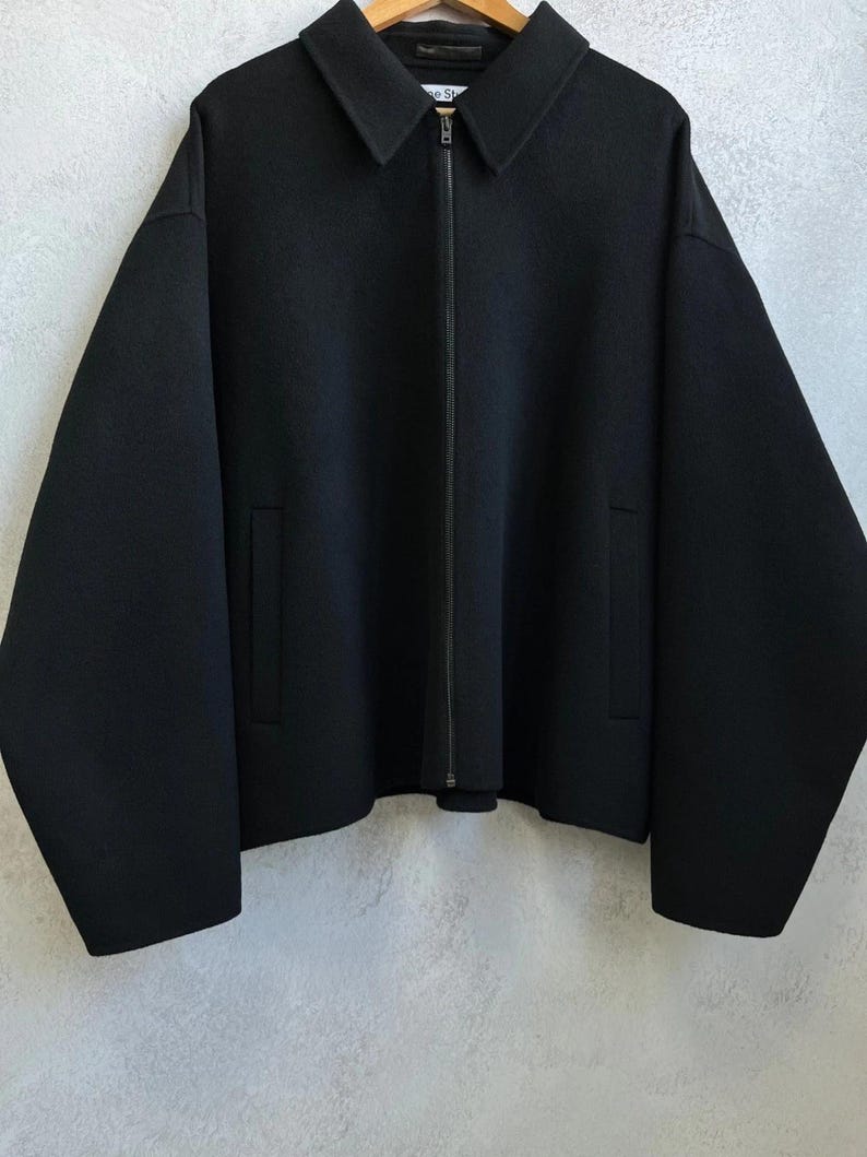 May include: A solid black jacket featuring a pointed collar, a full-length front zipper closure, and two vertical slit pockets. The garment has long, wide sleeves and a straight hemline, constructed from a smooth, dense fabric material.