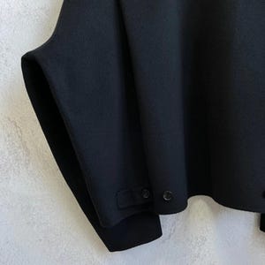 May include: A black jacket featuring a smooth, solid-colored fabric with a wide, structured sleeve design. The garment includes a horizontal seam across the upper back and a small tab detail at the lower hem secured with a single black button.