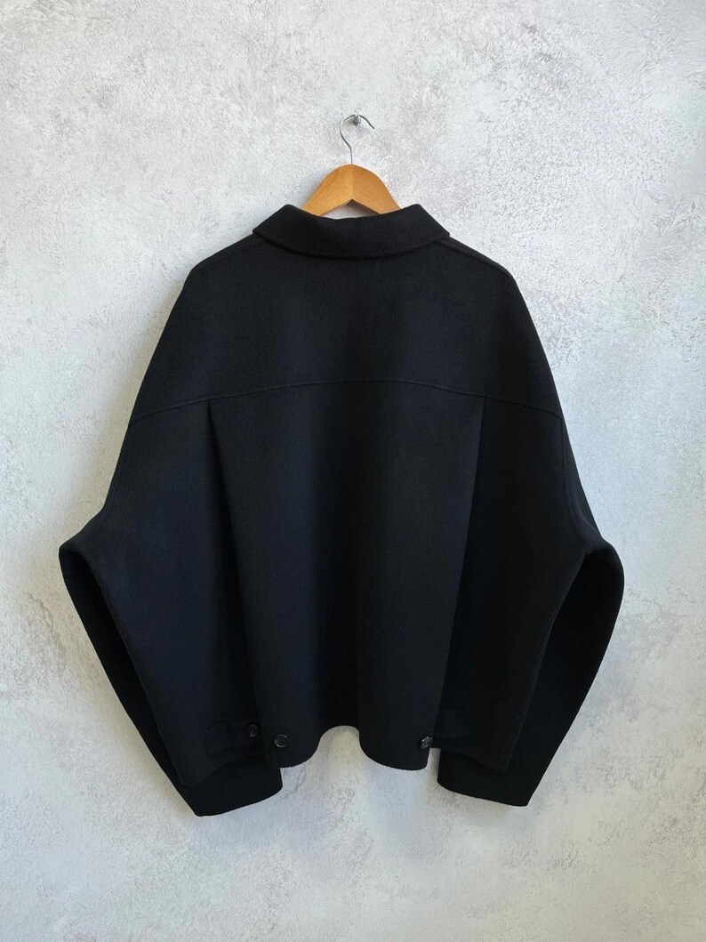 May include: A solid black jacket with a wide, boxy silhouette, a classic collar, and dropped shoulders. The garment features a horizontal back yoke, long sleeves, and a straight hemline with small button details at the lower sides for adjustment.