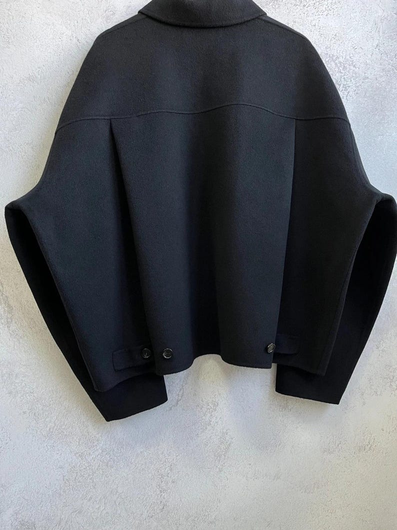 May include: A black jacket with a wide, boxy silhouette, featuring a classic collar, dropped shoulders, and long sleeves. The back displays a horizontal yoke seam, vertical pleats, and two small tabs at the hem, each secured with a single black button.