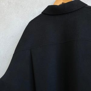 May include: A solid black jacket featuring a classic collar, a horizontal seam across the upper back, and a textured fabric finish. The garment is displayed on a wooden hanger, showcasing a structured design with clean lines and a minimalist aesthetic.