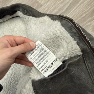 May include: A charcoal gray jacket with a white faux shearling lining and a metal zipper. A white fabric care label is attached to the interior, featuring the text Acne Studios and detailed composition information in multiple languages.