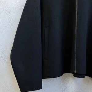 May include: A solid black jacket featuring a full-length front zipper closure, long sleeves, and a vertical welt pocket on the front panel. The garment is constructed from a smooth, dense fabric with a clean, minimalist aesthetic and straight hem.