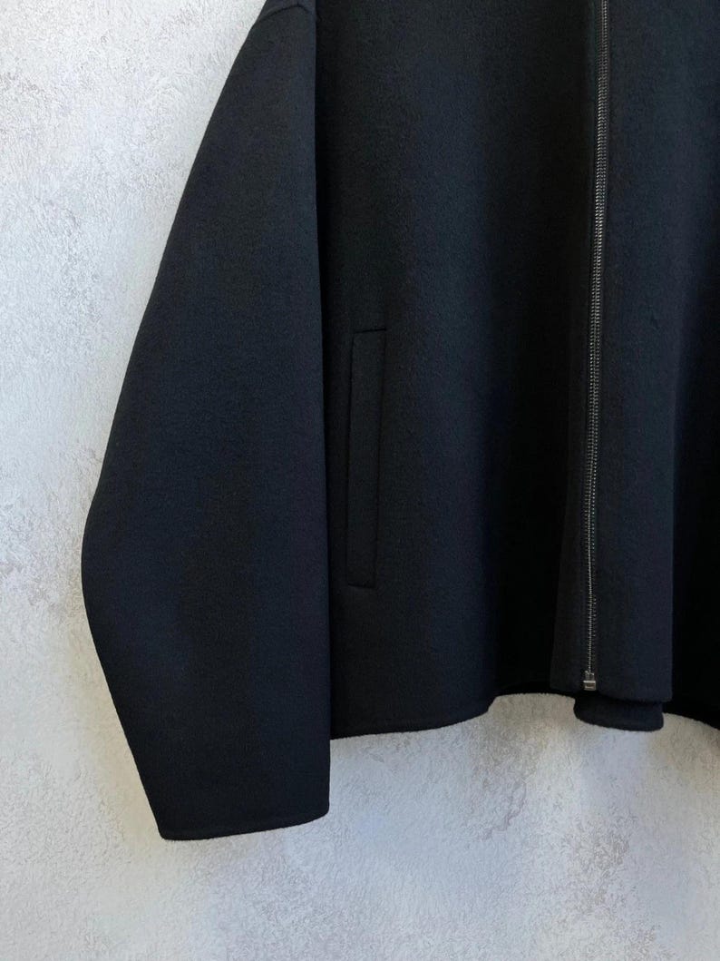 May include: A solid black jacket featuring a full-length front zipper closure, long sleeves, and a vertical welt pocket on the front panel. The garment is constructed from a smooth, dense fabric with a clean, minimalist aesthetic and straight hem.