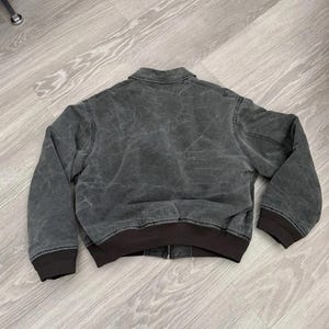 May include: A charcoal gray jacket with a distressed, washed-out texture, featuring a classic fold-down collar, long sleeves with dark brown ribbed cuffs, and a matching dark brown ribbed hem. The jacket includes a central front zipper closure.