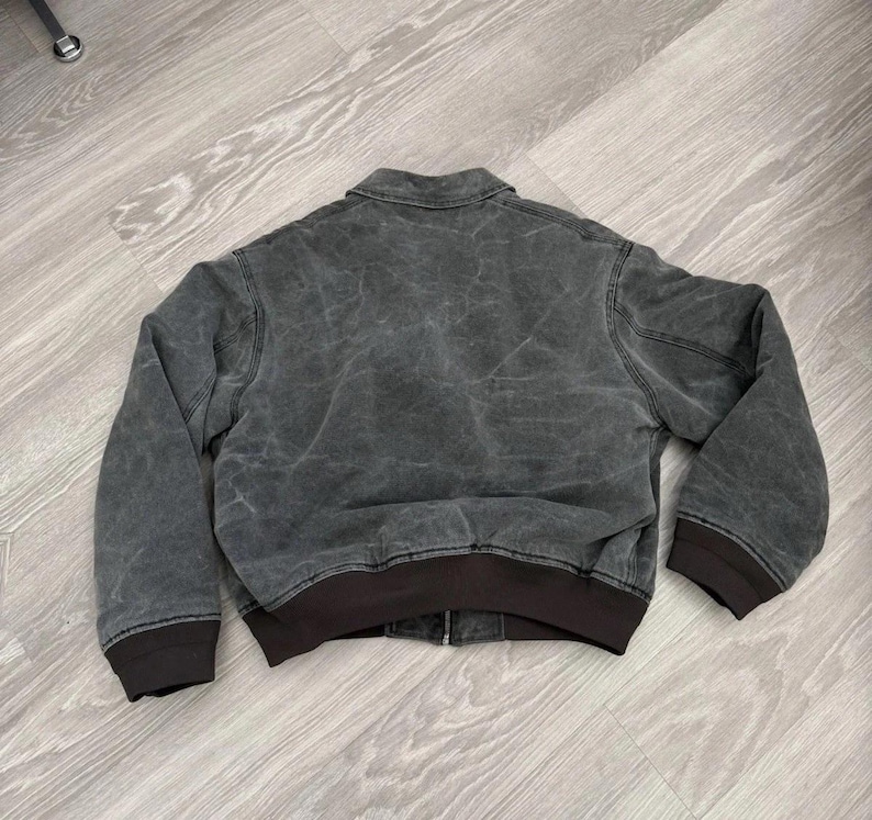 May include: A charcoal gray jacket with a distressed, washed-out texture, featuring a classic fold-down collar, long sleeves with dark brown ribbed cuffs, and a matching dark brown ribbed hem. The jacket includes a central front zipper closure.