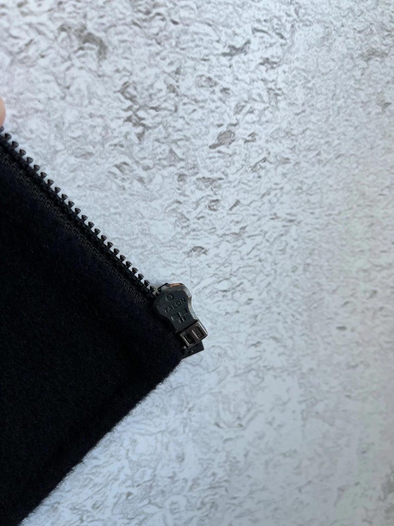 May include: A black fabric garment edge featuring a black metal zipper track and a matching black metal zipper pull. The zipper pull is embossed with the word TALON and a small circular logo, showing a functional closure mechanism for clothing.