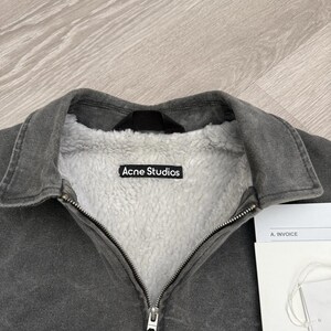 May include: A charcoal gray jacket with a white sherpa fleece lining and a fold-over collar. The interior features a black label with the white text Acne Studios. A metal zipper runs down the center, and a white invoice document is visible nearby.