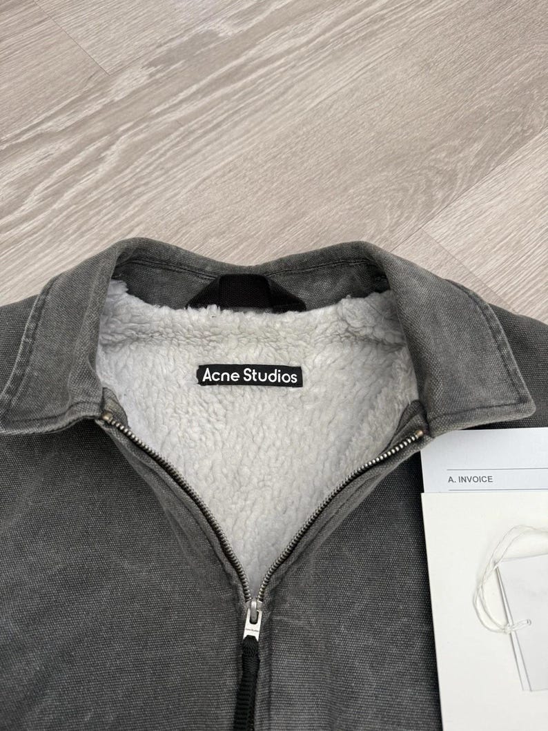 May include: A charcoal gray jacket with a white sherpa fleece lining and a fold-over collar. The interior features a black label with the white text Acne Studios. A metal zipper runs down the center, and a white invoice document is visible nearby.