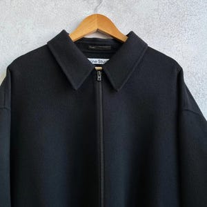 May include: A black zip-up jacket with a pointed collar and a full-length front zipper. The interior neck label features the text Acne Studios. The garment is constructed from a solid black fabric with a smooth texture and long sleeves.