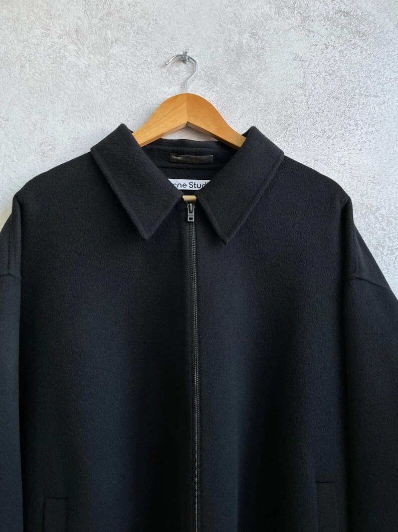 May include: A black zip-up jacket with a pointed collar and a full-length front zipper. The interior neck label features the text Acne Studios. The garment is constructed from a solid black fabric with a smooth texture and long sleeves.