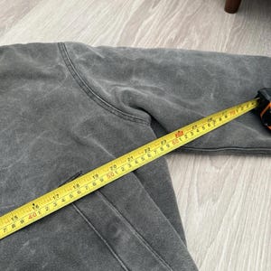 May include: A charcoal gray jacket with a visible seam and a pocket, measured by a yellow tape measure showing a length of approximately 28 inches from the shoulder area to the sleeve cuff, indicating the garment's scale in a clear, objective view.