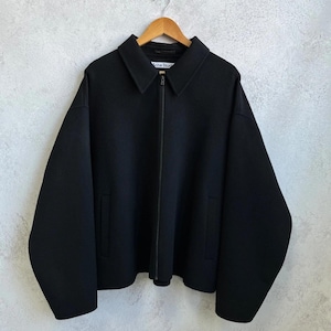 May include: A black zip-up jacket with a pointed collar, long sleeves, and two side pockets. The garment features a central zipper closure and a white interior label with the text Acne Studios, displayed on a wooden hanger against a textured wall.