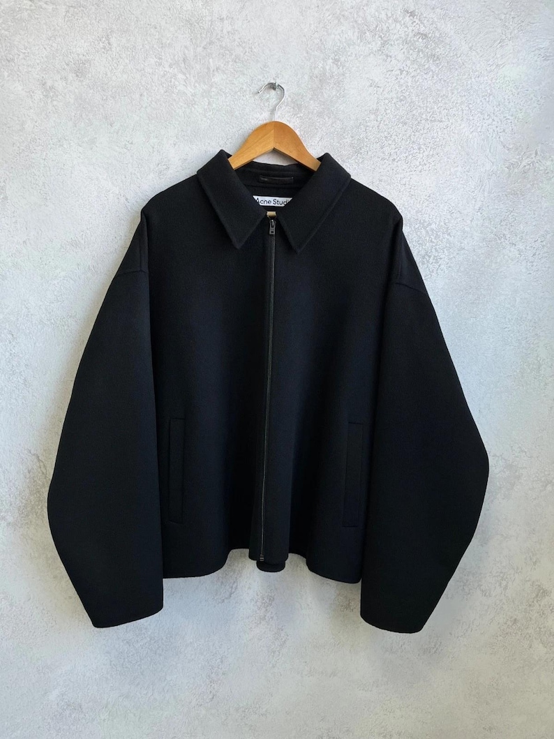 May include: A black zip-up jacket with a pointed collar, long sleeves, and two side pockets. The garment features a central zipper closure and a white interior label with the text Acne Studios, displayed on a wooden hanger against a textured wall.