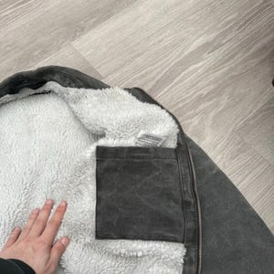 May include: A charcoal gray jacket featuring a thick white sherpa fleece lining. The interior includes a square patch pocket made of the same charcoal gray fabric as the exterior shell, along with a white care label tag near the collar.