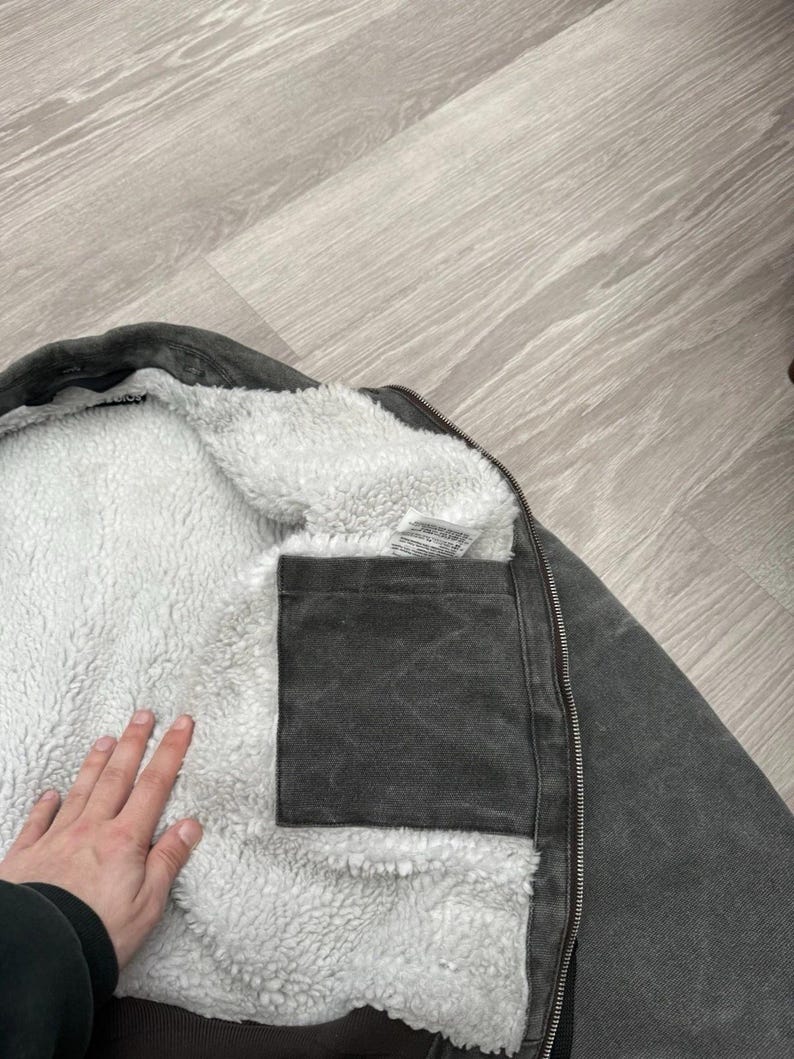 May include: A charcoal gray jacket featuring a thick white sherpa fleece lining. The interior includes a square patch pocket made of the same charcoal gray fabric as the exterior shell, along with a white care label tag near the collar.