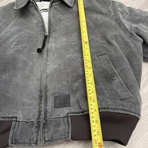 May include: A charcoal gray zip-up jacket with a white faux-fur lining, a collar, and a black leather patch featuring a face icon. A yellow measuring tape is placed vertically along the front, showing a length of approximately 25 inches.