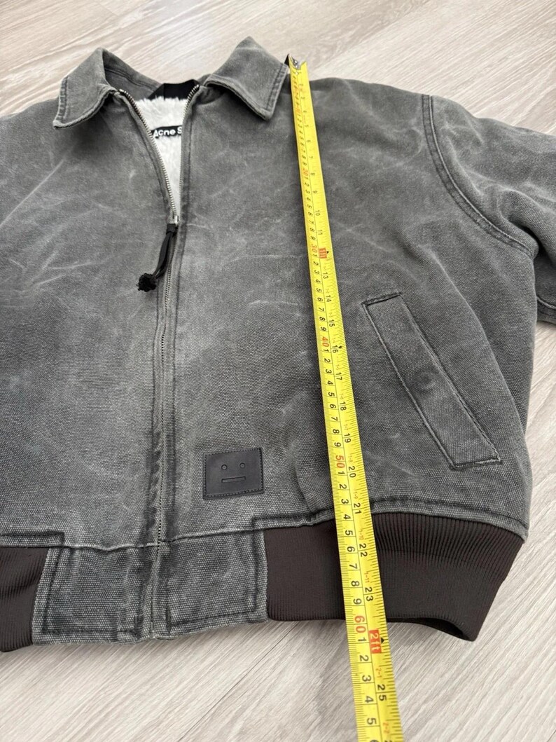 May include: A charcoal gray zip-up jacket with a white faux-fur lining, a collar, and a black leather patch featuring a face icon. A yellow measuring tape is placed vertically along the front, showing a length of approximately 25 inches.
