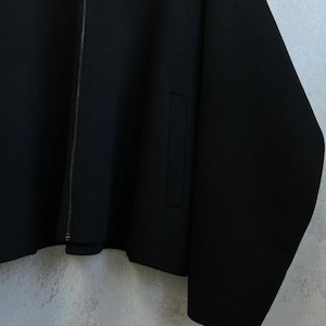 May include: A solid black jacket featuring a full-length vertical zipper closure down the center. The garment includes long sleeves and a single visible vertical slit pocket on the front side. The fabric appears to have a smooth, uniform texture.