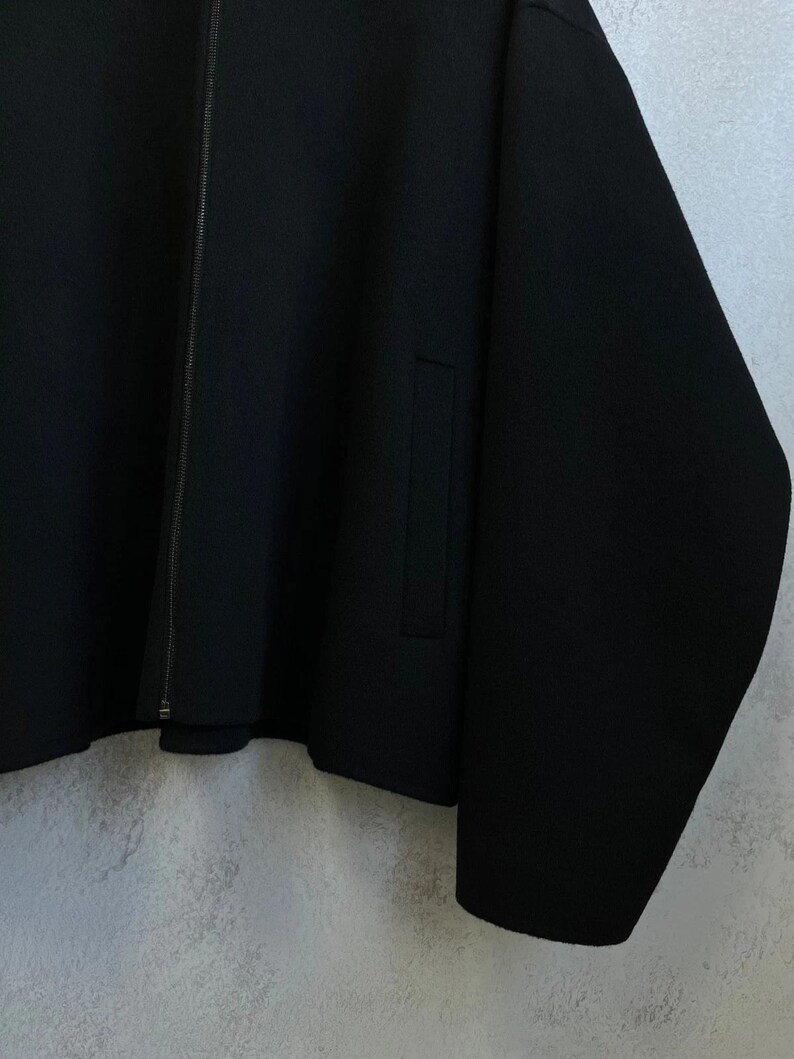 May include: A solid black jacket featuring a full-length vertical zipper closure down the center. The garment includes long sleeves and a single visible vertical slit pocket on the front side. The fabric appears to have a smooth, uniform texture.