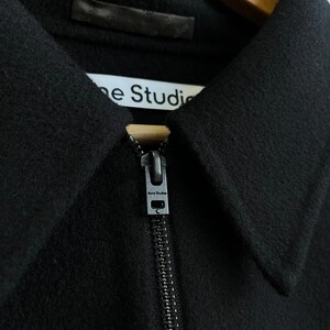 May include: A black jacket with a pointed collar and a full-length front zipper. The zipper pull is metallic and features the text Acne Studios. A white interior label is visible at the neckline with the text Acne Studios printed in black.