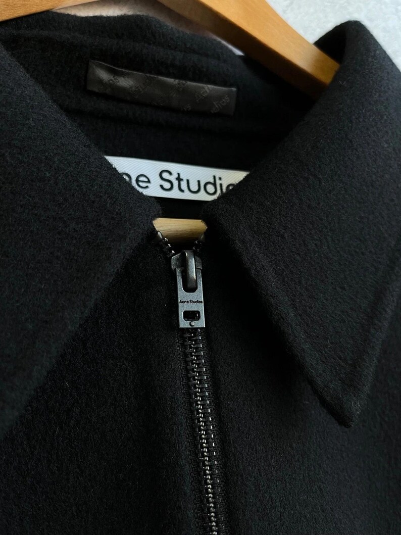 May include: A black jacket with a pointed collar and a full-length front zipper. The zipper pull is metallic and features the text Acne Studios. A white interior label is visible at the neckline with the text Acne Studios printed in black.