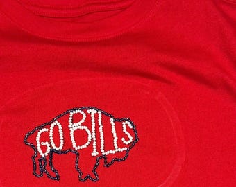 May include: A bright red t-shirt featuring a beaded graphic of a buffalo silhouette. The outline of the buffalo is created with dark blue beads, while the interior text reads GO BILLS in white beads, centered on the chest of the garment.