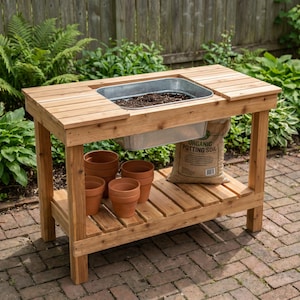 Pode incluir: Uma bancada de jardinagem de madeira com uma bacia retangular de metal galvanizado embutida no tampo. A bancada inclui uma prateleira inferior com ripas contendo vários vasos de terracota e um saco de terra orgânica com texto visível.