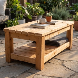 May include: A rectangular wooden coffee table with a slatted top and a lower shelf. The table is constructed from light brown wood planks. A grey folded blanket rests on the lower shelf, while a ceramic mug and a book sit on the top surface.